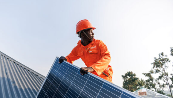 Amped Innovation worker wearing hi-viz orange branded jumpsuit and matching hardhat holding solar panel during rooftop installation, looking into the distance.