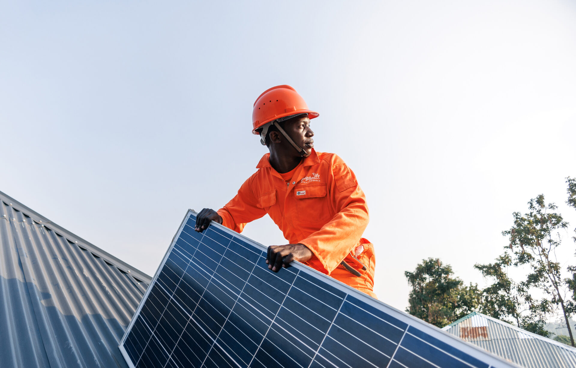 Amped Innovation worker wearing hi-viz orange branded jumpsuit and matching hardhat holding solar panel during rooftop installation, looking into the distance.