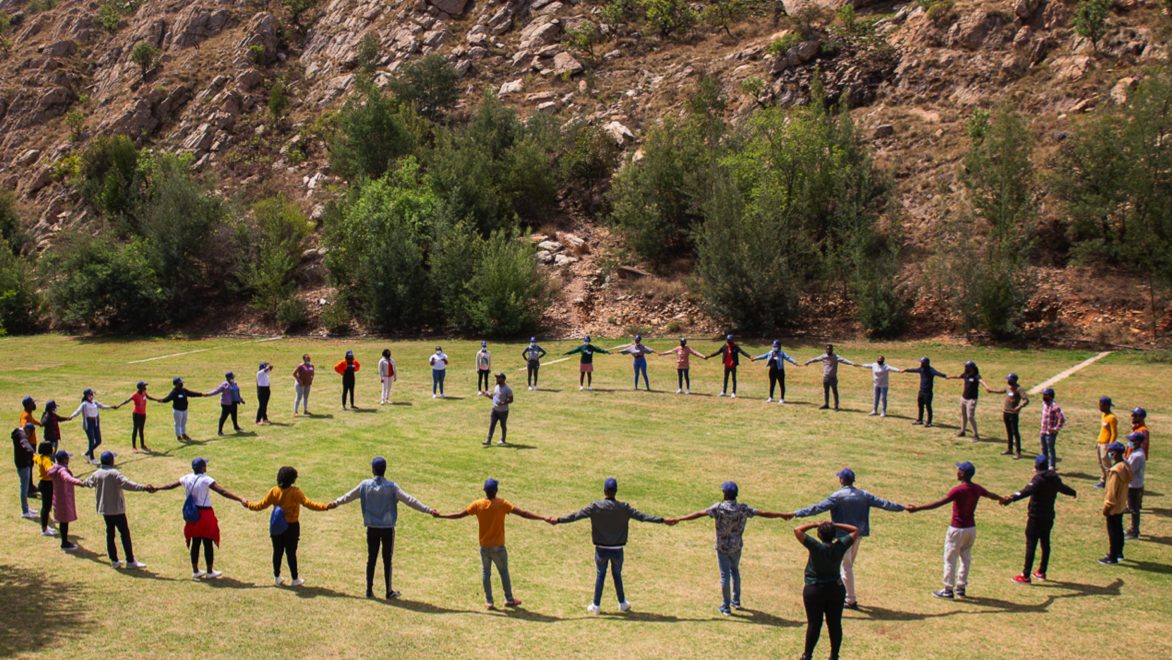 A group of a few dozen South African artisan apprentices participating in a BluLever Education training exercise , all holding hands to form a large circle on a grassy field on a sunny day, with a facilitator in the center. Background: young trees and shrubs on a sloped, rocky hillside.