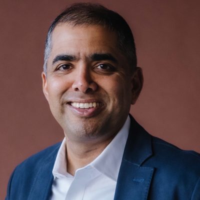 Headshot of Rahul Shendure, CarbonBuilt CEO — smiling and looking at the camera, wearing a white collared shirt and a blue blazer