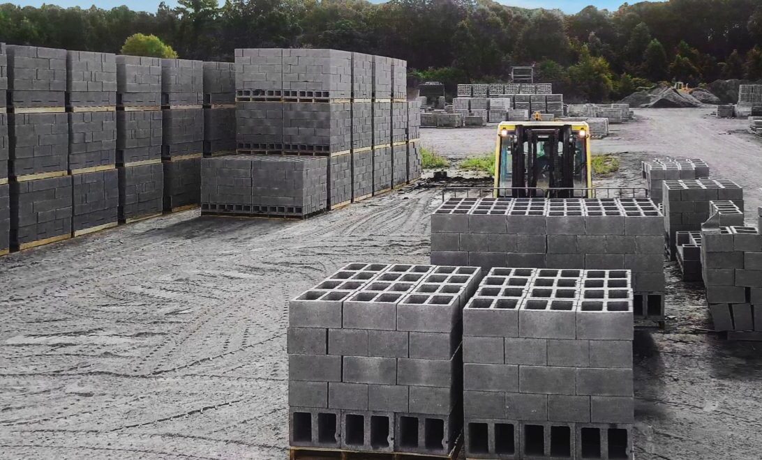 Wide angle view of dozens of pallets of concrete blocks in an open-air dirt field on an overcast day. Foreground: A person in a forklift facing the camera, behind a few large pallets of concrete blocks.