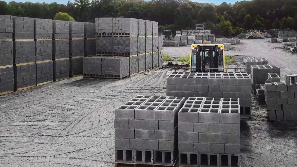 Wide angle view of dozens of pallets of concrete blocks in an open-air dirt field on an overcast day. Foreground: A person in a forklift facing the camera, behind a few large pallets of concrete blocks.