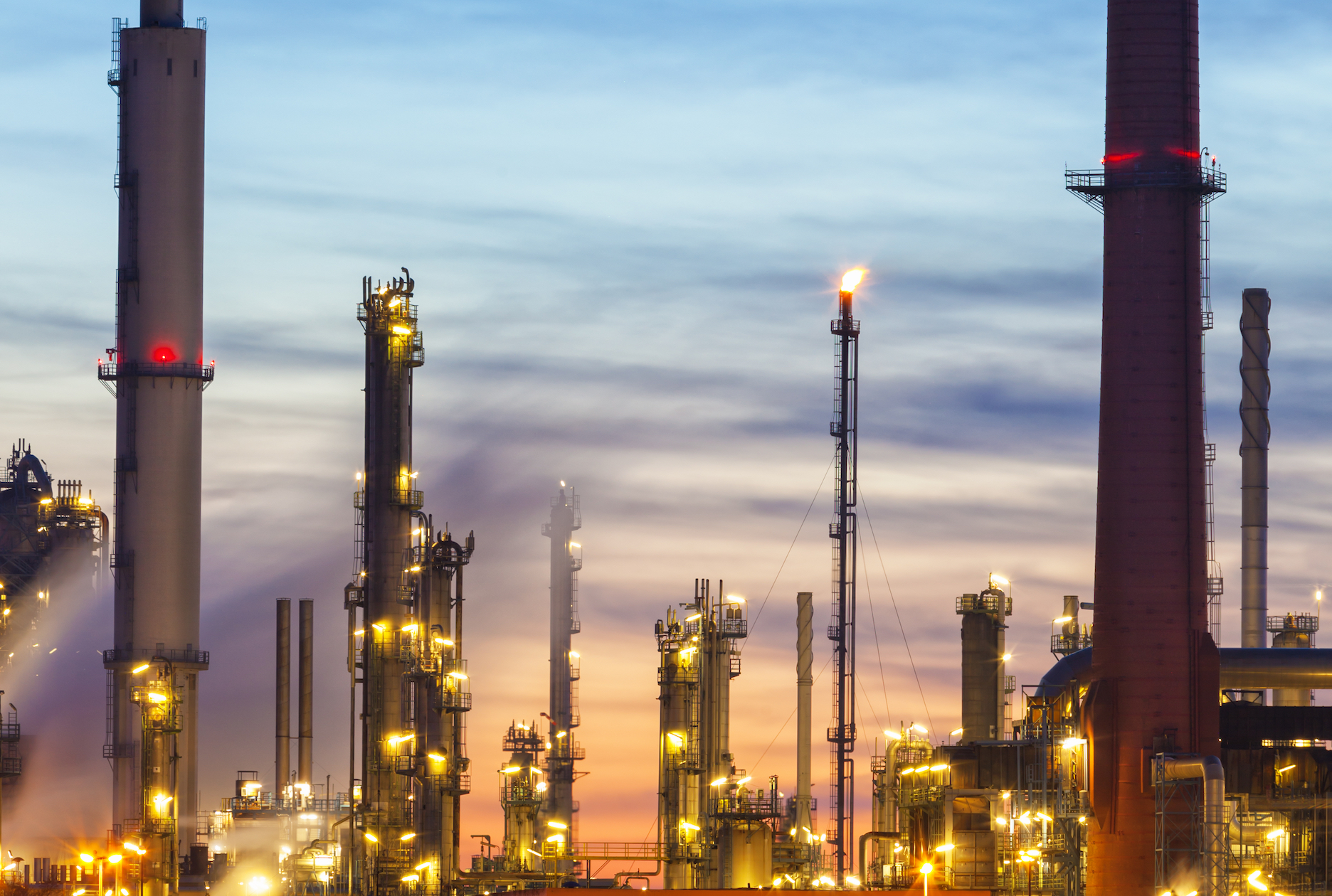 Skyline view of oil refinery in Germany, illuminated at dusk. Natural gas flaring out of one of numerous smokestacks.