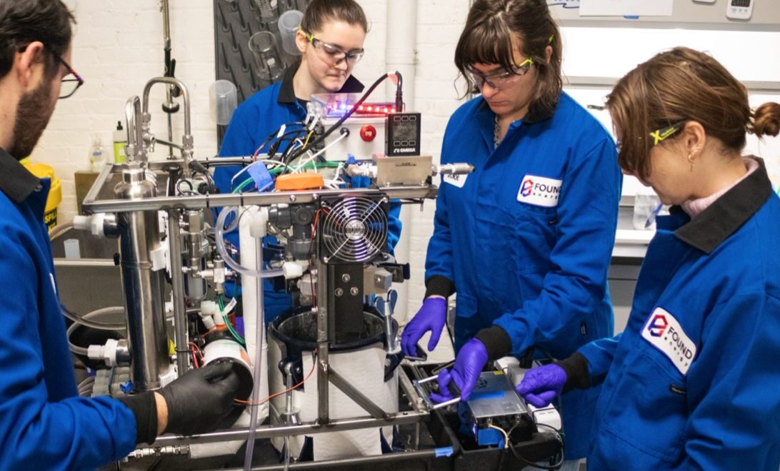 Four Found Energy employees&mdash;1 man (far-left), three women (middle, middle-right, right) wearing blue Found Energy branded jumpsuits with black collars and protective eyewear and latex gloves. The group is standing around, working on, and looking at a contraption with metal rods, electric wiring, mechanical components, and a small computer fan. Setting: Fluorescent-it lab space.