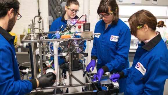 Four Found Energy employees&mdash;1 man (far-left), three women (middle, middle-right, right) wearing blue Found Energy branded jumpsuits with black collars and protective eyewear and latex gloves. The group is standing around, working on, and looking at a contraption with metal rods, electric wiring, mechanical components, and a small computer fan. Setting: Fluorescent-it lab space.