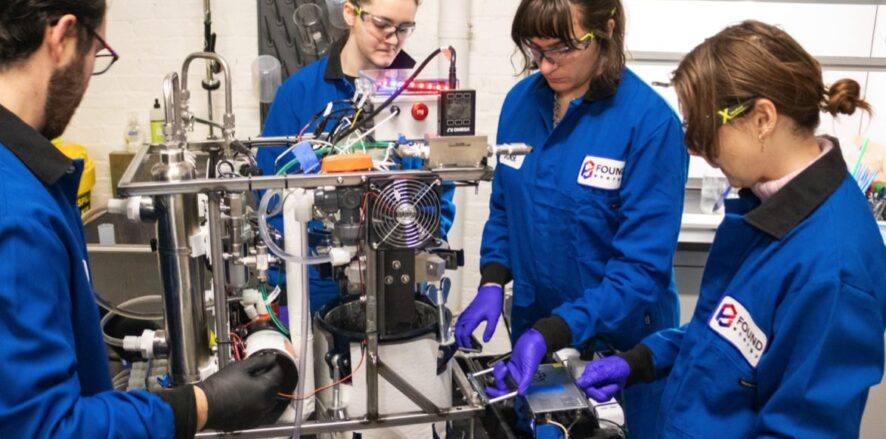 Four Found Energy employees—1 man (far-left), three women (middle, middle-right, right) wearing blue Found Energy branded jumpsuits with black collars and protective eyewear and latex gloves. The group is standing around, working on, and looking at a contraption with metal rods, electric wiring, mechanical components, and a small computer fan. Setting: Fluorescent-it lab space.