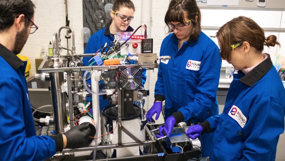 Four Found Energy employees—1 man (far-left), three women (middle, middle-right, right) wearing blue Found Energy branded jumpsuits with black collars and protective eyewear and latex gloves. The group is standing around, working on, and looking at a contraption with metal rods, electric wiring, mechanical components, and a small computer fan. Setting: Fluorescent-it lab space.