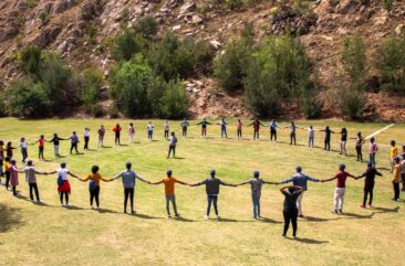 A group of a few dozen South African artisan apprentices participating in a BluLever Education training exercise , all holding hands to form a large circle on a grassy field on a sunny day, with a facilitator in the center. Background: young trees and shrubs on a sloped, rocky hillside.