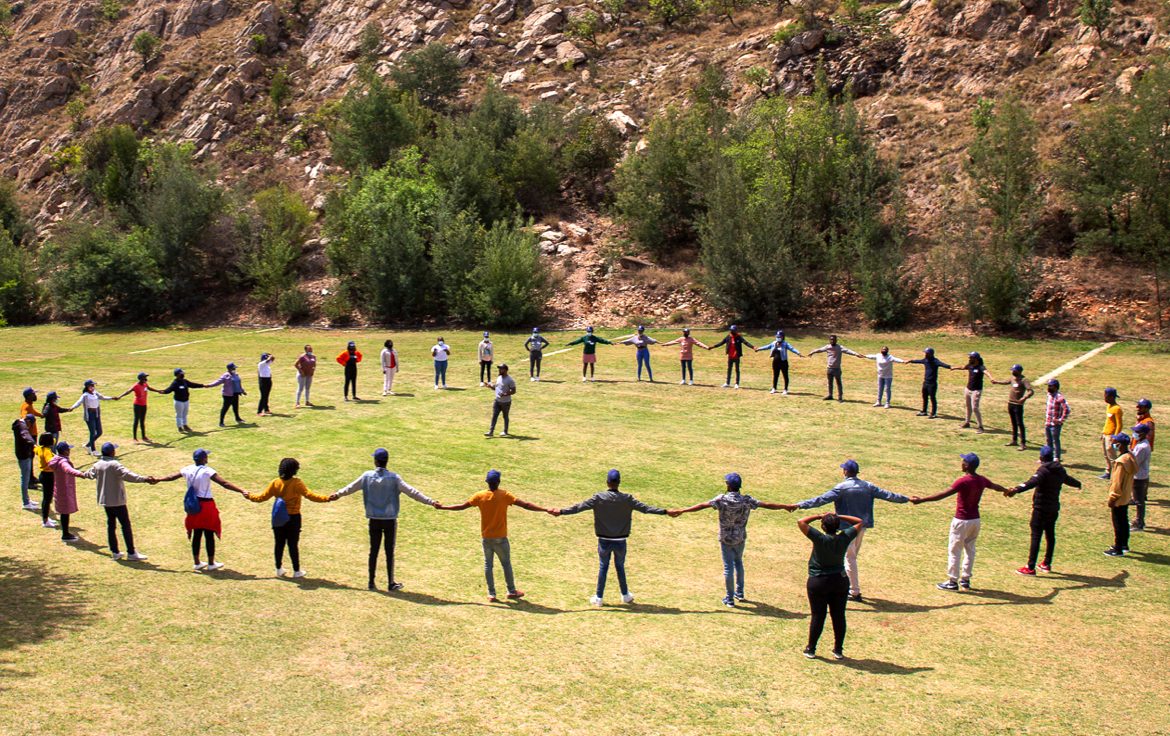 A group of a few dozen South African artisan apprentices participating in a BluLever Education training exercise , all holding hands to form a large circle on a grassy field on a sunny day, with a facilitator in the center. Background: young trees and shrubs on a sloped, rocky hillside.