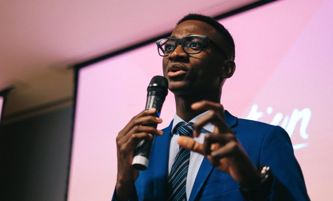 Close-up candid/action shot of a young man wearing a blue blazer, glasses, button-down shirt and a striped blue tie, holding a microphone in his right hand and his left hand in front of him, fingers curled as if grabbing something, presenting at a Generation workshop or event.