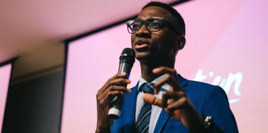 Close-up candid/action shot of a young man wearing a blue blazer, glasses, button-down shirt and a striped blue tie, holding a microphone in his right hand and his left hand in front of him, fingers curled as if grabbing something, presenting at a Generation workshop or event.