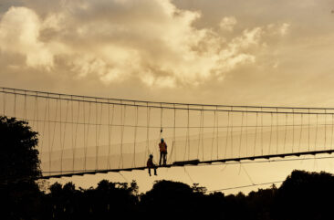 Two construction workers on a Bridges to Prosperity trailbridge—one sitting, one standing—at dusk, surrounded by trees.