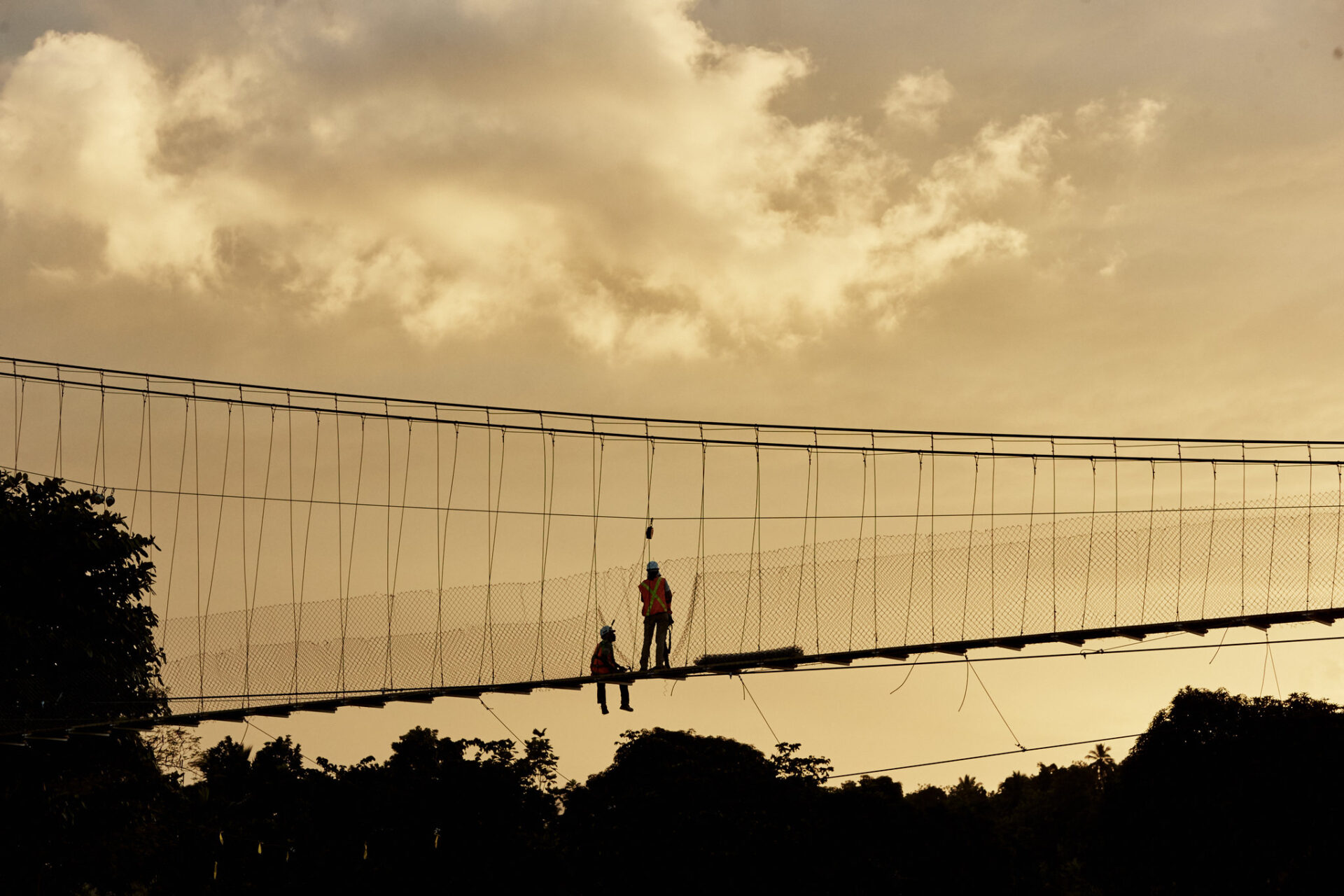 Two construction workers on a Bridges to Prosperity trailbridge—one sitting, one standing—at dusk, surrounded by trees.