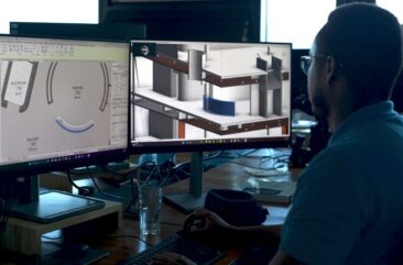 Over-the-left-shoulder view of MASS Design Group employee looking at building designs on two monitors, sitting at a desk. Individual is wearing glasses and a blue polo, with his right hand operating the mouse and his left hand resting on the keyboard.