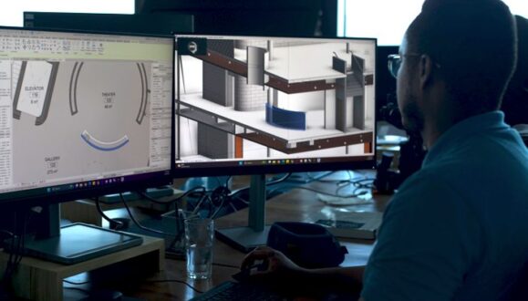 Over-the-left-shoulder view of MASS Design Group employee looking at building designs on two monitors, sitting at a desk. Individual is wearing glasses and a blue polo, with his right hand operating the mouse and his left hand resting on the keyboard.