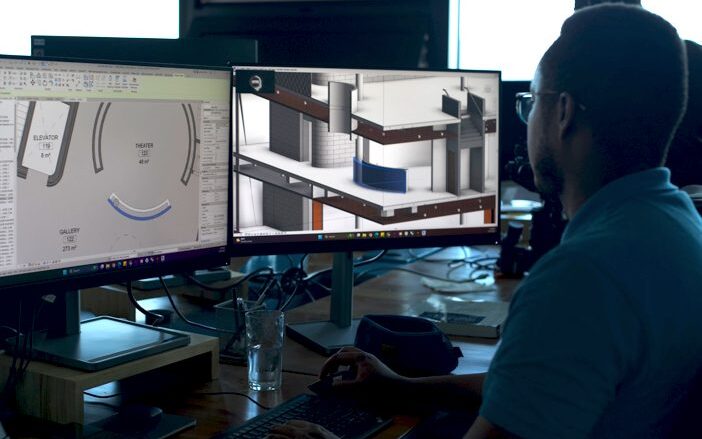 Over-the-left-shoulder view of MASS Design Group employee looking at building designs on two monitors, sitting at a desk. Individual is wearing glasses and a blue polo, with his right hand operating the mouse and his left hand resting on the keyboard.