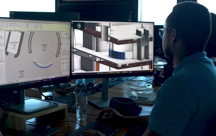 Over-the-left-shoulder view of MASS Design Group employee looking at building designs on two monitors, sitting at a desk. Individual is wearing glasses and a blue polo, with his right hand operating the mouse and his left hand resting on the keyboard.