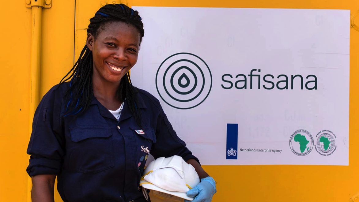 A woman Safisana employee standing in front of a Safisana sign at the Ashaiman recycling plant, smiling and looking at the camera, holding a white hard hat in her blue-gloved left hand.