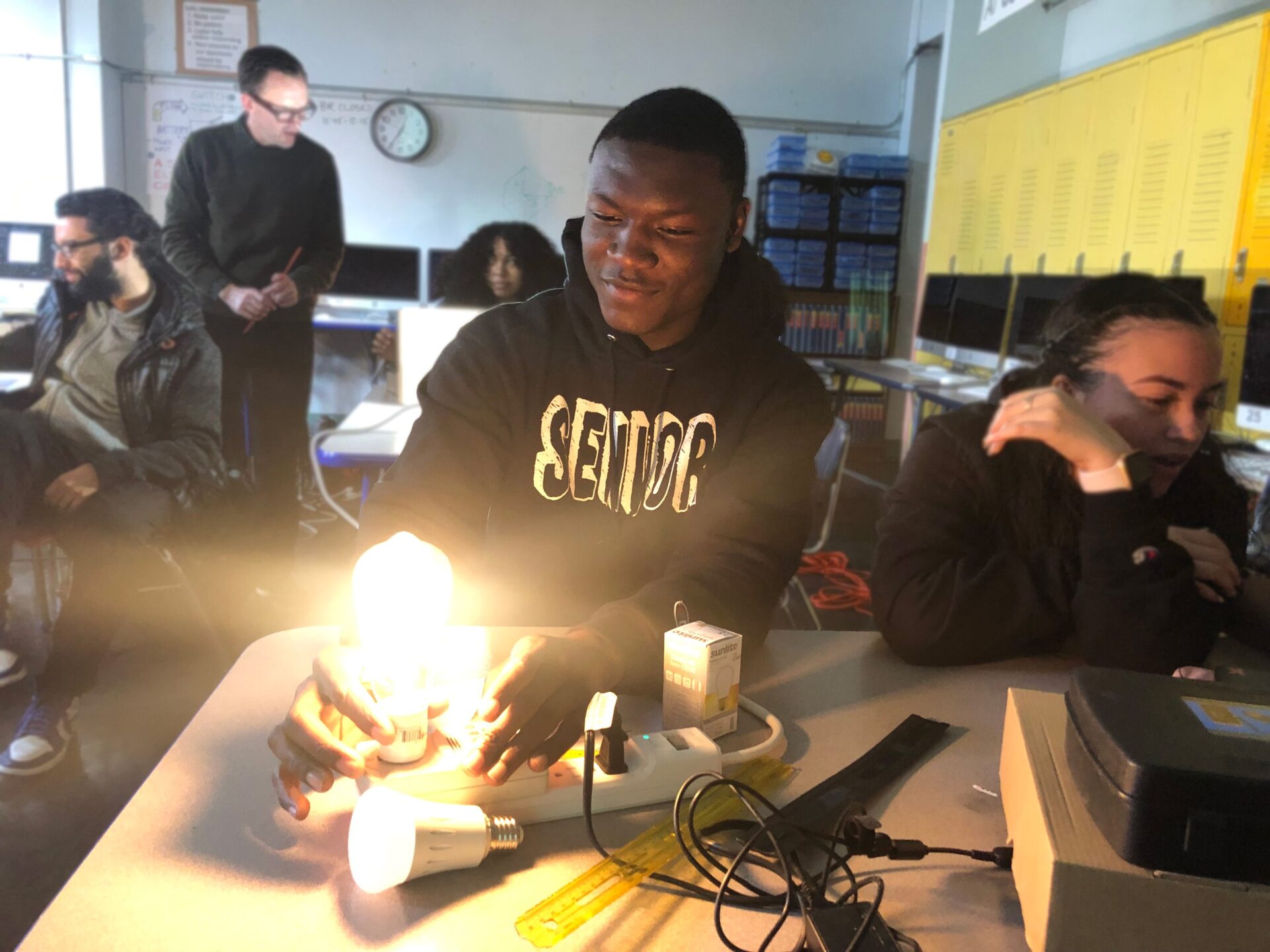 Student wearing "Senior" hoodie, smiling while experimenting with light project in a classroom with other students sitting at desks/tables and instructor standing in the background.