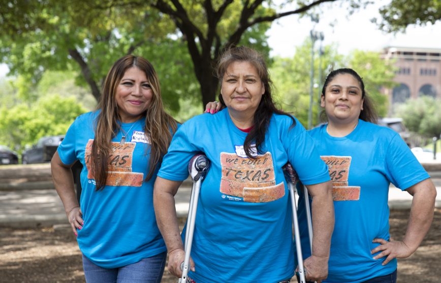 Families and workers from Better Builder (FWF grantee) in Austin, Texas — Three women standing side-by-side, smiling and posing for group photo, wearing matching blue "We Build Texas" t-shirts and adhesive name-tags. The woman in the middle is leaning on a pair of crutches.