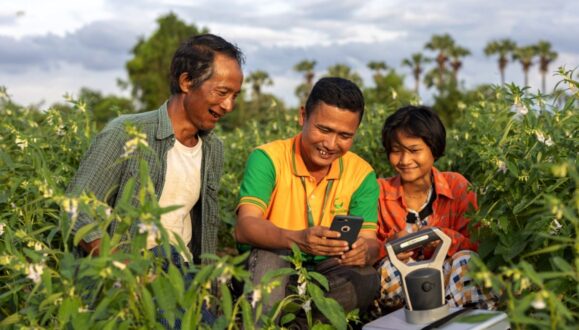 Three individuals—an older man, a middle-aged man, and a teenager—standing in a farm field surrounded by tall green crops on a sunny day. All three are smiling and looking at a mobile phone, held by the middle-aged man, which appears to interacting with a sensor or other mechanism for monitoring the crop.