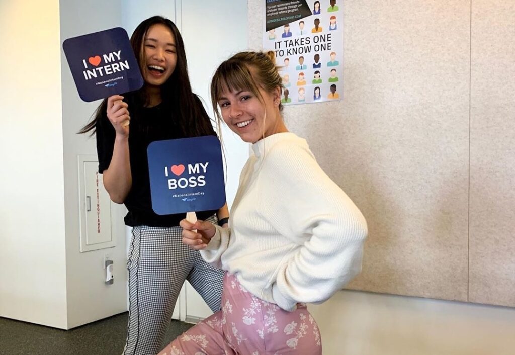 An Autodesk intern holding a sign that says “I heart my boss” stands to the right of her manager, who is holding a sign that says “I heart my intern.”