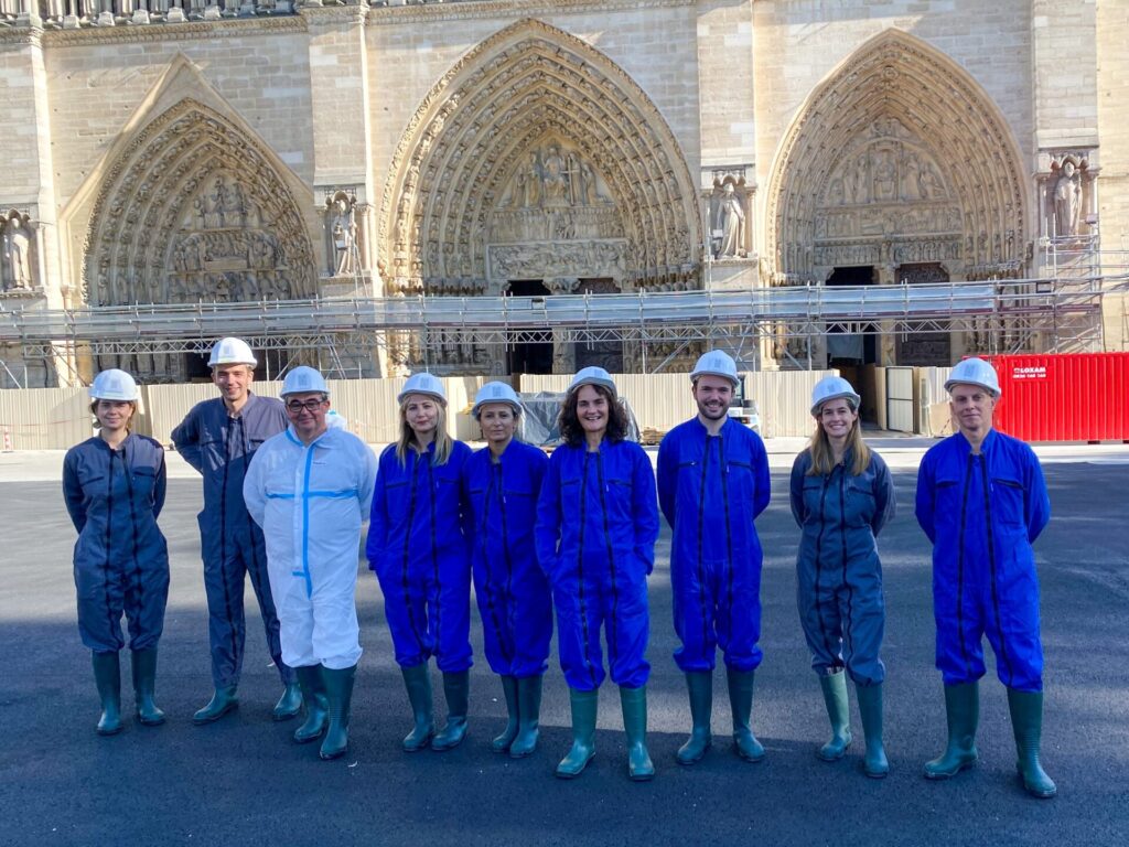 A group of Autodesk employees stand in front of the Notre Dame cathedral in Paris wearing protective clothing and white hard hats