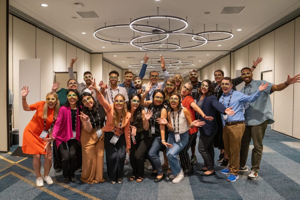 A large group of Autodesk's employee resource group (ERG) members stands in a large room at a convention center. Most of them have their hands up in the air with happy expressions. and are wearing Mardi Gras-style masks.