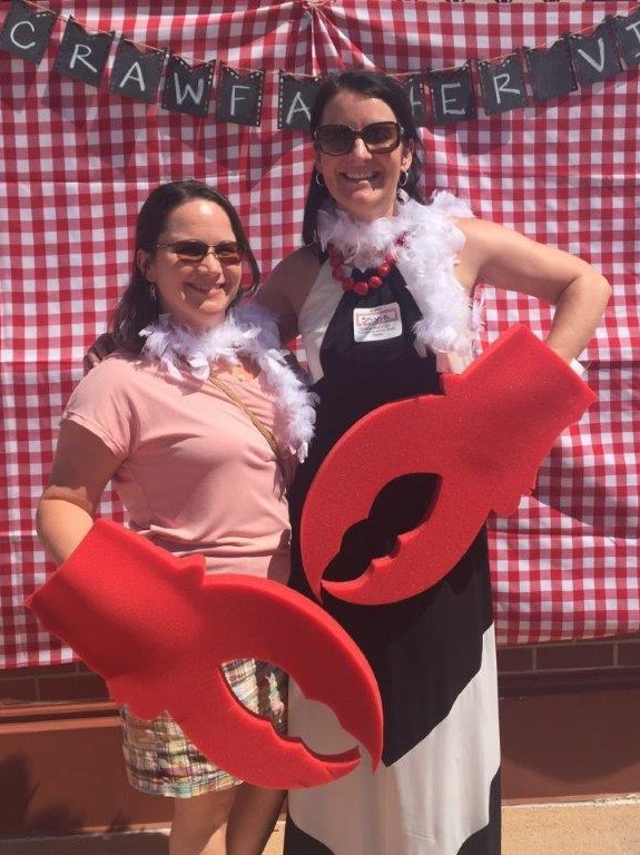 At a "crawfather" fundraiser, Colleen Lucksinger and Shona McMoran stand in front of a red and white plaid backdrop with large foam lobster claws on their hands.