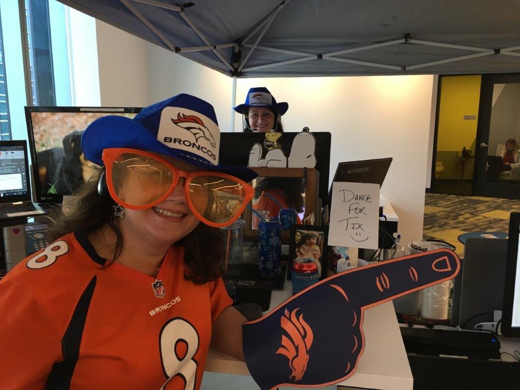 Colleen Lucksinger and Shona McMoran stand at their desks wearing Denver Broncos hats and jerseys for a sports-themed Call Out Day in the office. Colleen is wearing oversized orange glasses and holds up a foam finger.