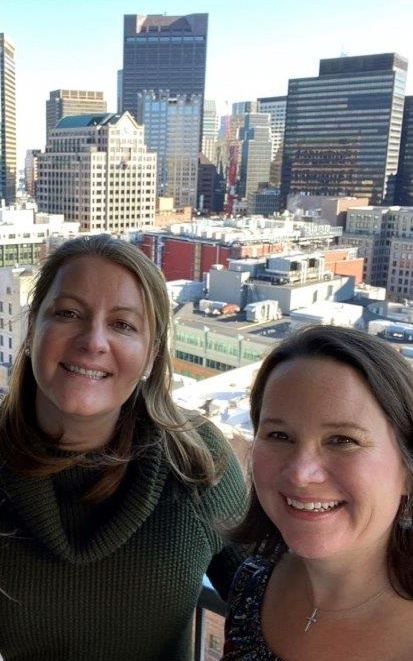 Shona McMoran and Colleen Lucksinger stand on a balcony with the city of Boston in the background.