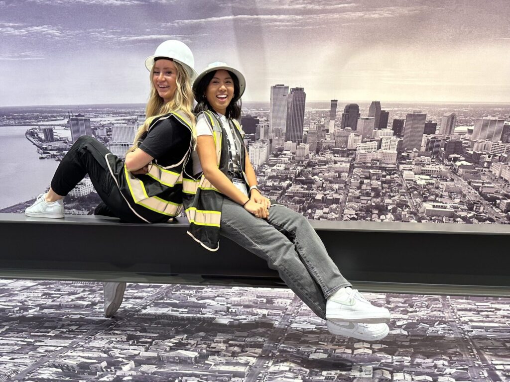 Kassidi Sorensen and Querida Xie sit on a metal beam in front of a mural of a large city, wearing white hard hats and construction vests