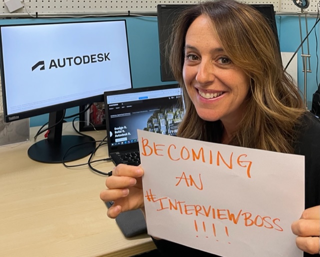 A female recruiter sits at a desk with a computer, holding up a sign that says “becoming a hashtag interview boss"