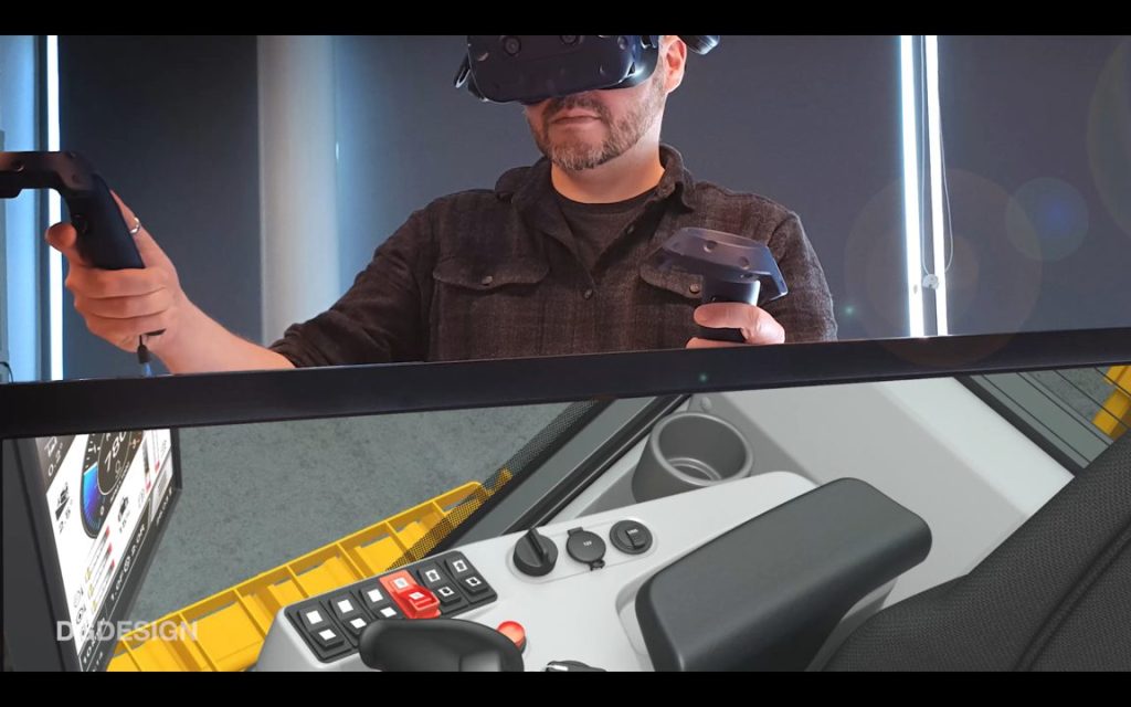 Top half of frame is a white man in VR goggles moving VR hand controls. Bottom half of frame is interior view of a bulldozer cab and control panels.