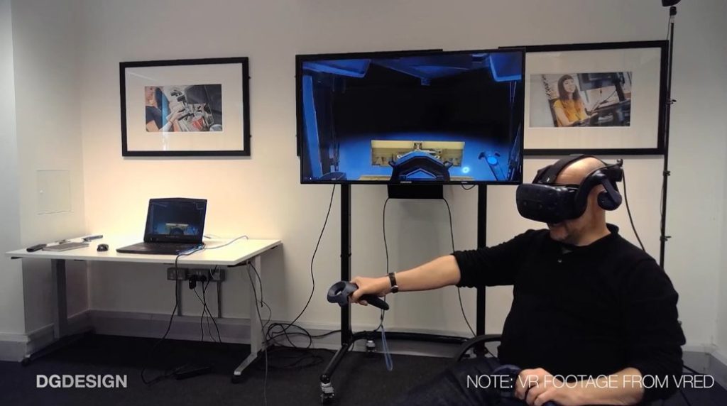 White man sitting down, computer monitor behind him. He's wearing VR goggles and using VR hand controls.