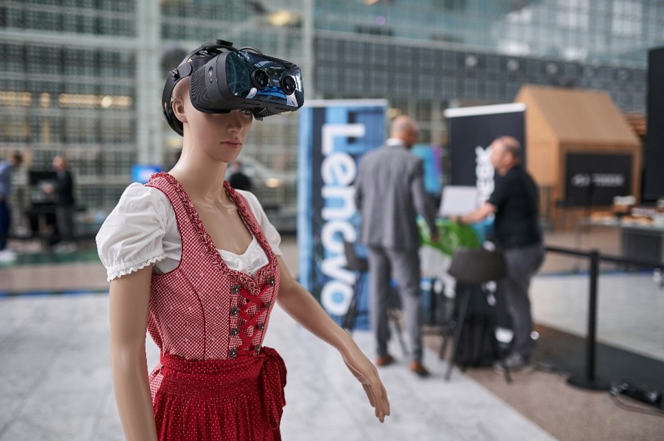 Woman in German outfit (red laced vest and white peasant blouse, red skirt) with VR goggles on. She's standing in a lobby with people in the background.