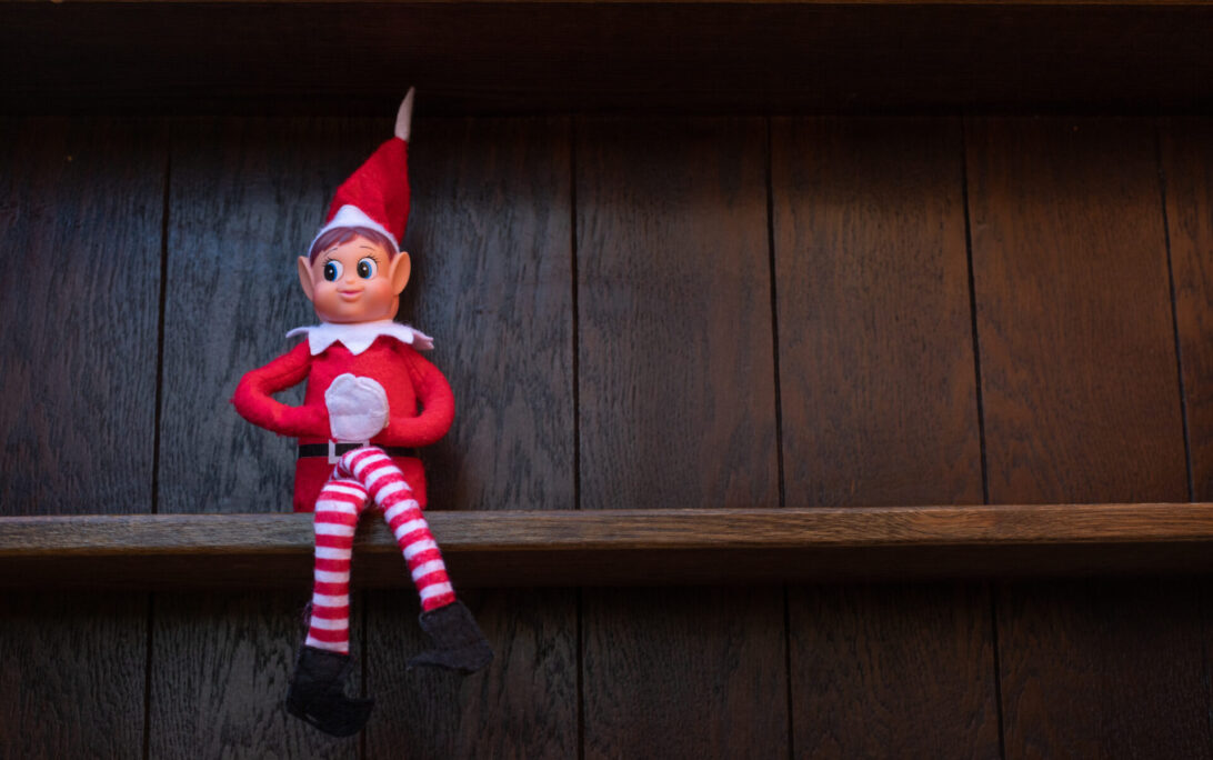 A small Christmas elf dressed in red and white, known as the Elf on the Shelf, is sitting on a dark wooden shelf.