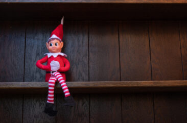 A small Christmas elf dressed in red and white, known as the Elf on the Shelf, is sitting on a dark wooden shelf.