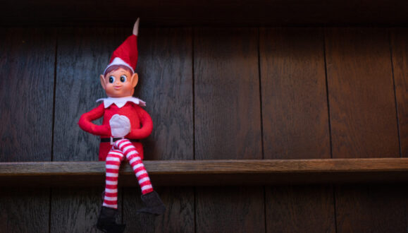 A small Christmas elf dressed in red and white, known as the Elf on the Shelf, is sitting on a dark wooden shelf.