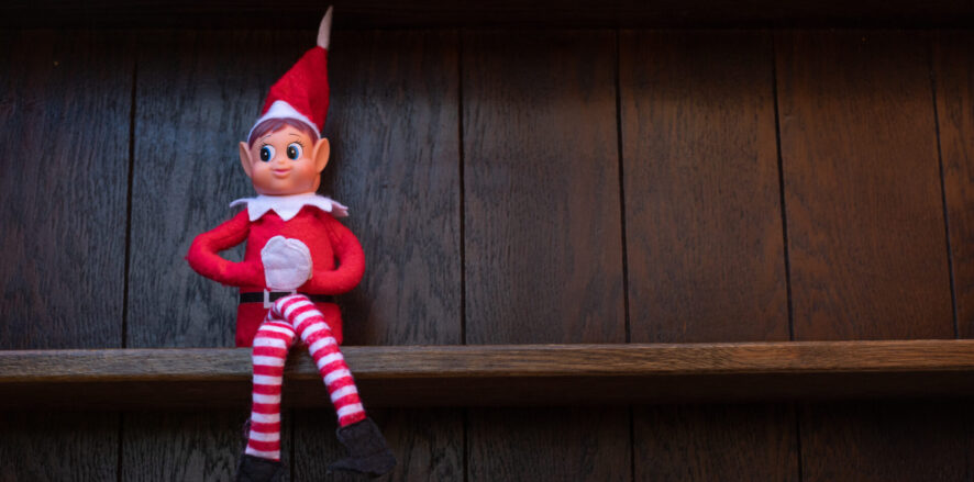 A small Christmas elf dressed in red and white, known as the Elf on the Shelf, is sitting on a dark wooden shelf.