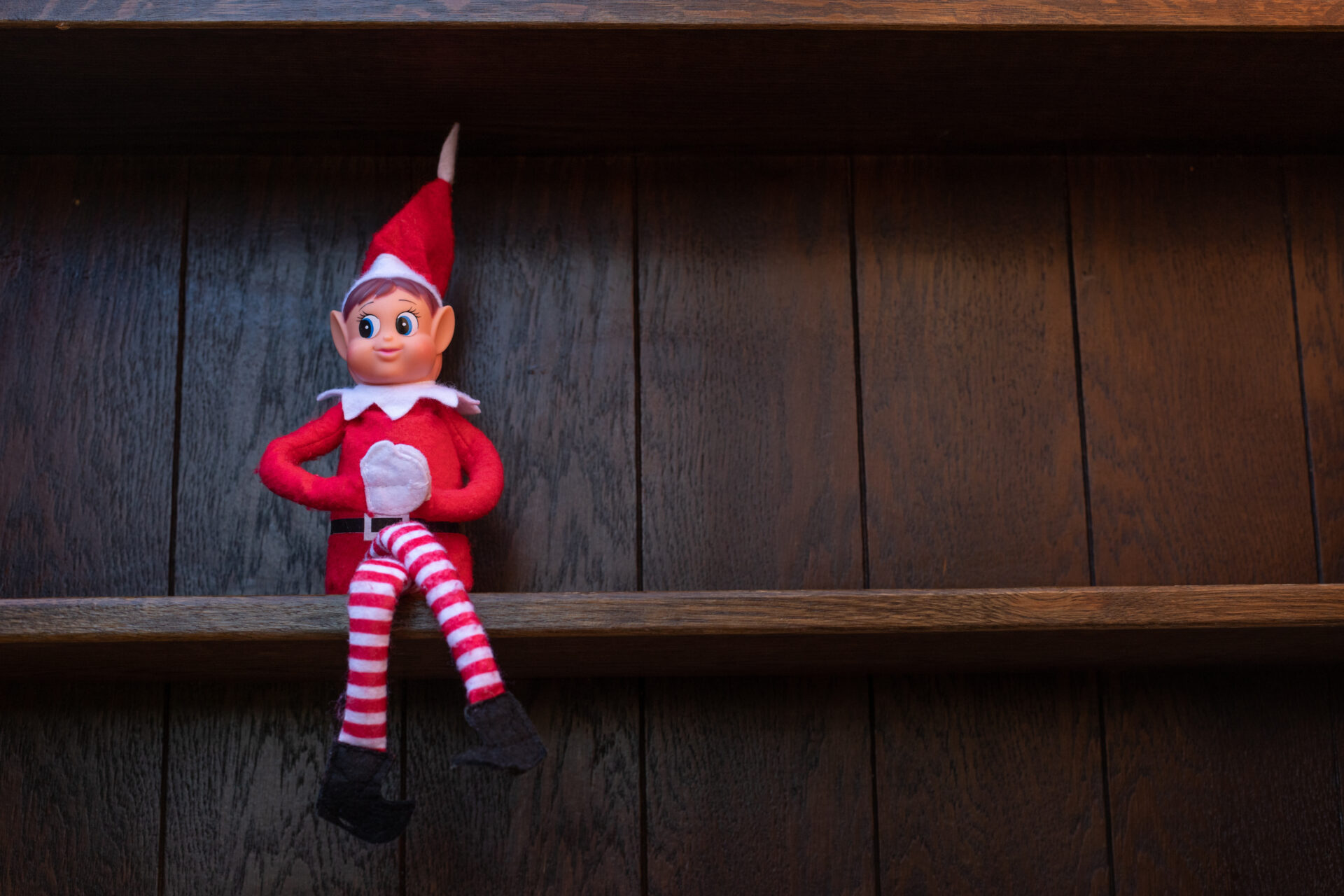 A small Christmas elf dressed in red and white, known as the Elf on the Shelf, is sitting on a dark wooden shelf.