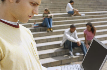Close-up of a college student working on his laptop on campus with other students in the background