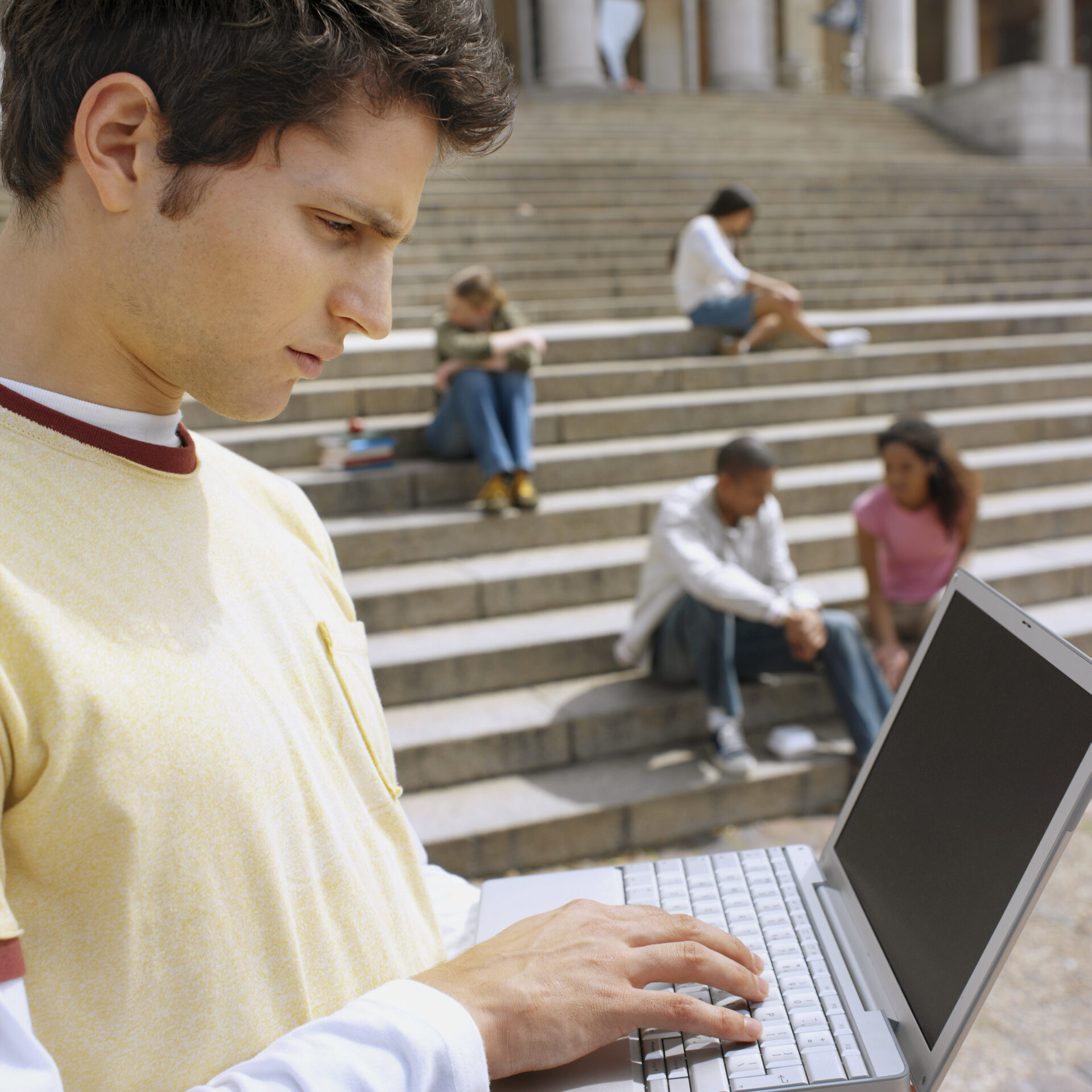 Close-up of a college student working on his laptop on campus with other students in the background